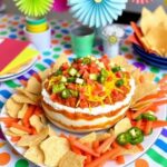 "Layered easy taco dip with creamy sour cream, refried beans, salsa, shredded cheese, and fresh toppings, served with tortilla chips for a party."